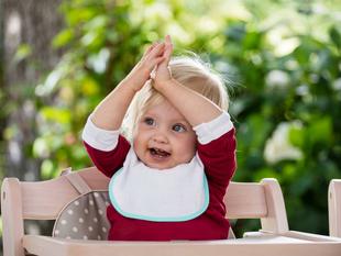 Ein kleines Kind mit blondem Haar, das ein rotes Hemd und ein weißes Lätzchen trägt, sitzt in einem hölzernen Hochstuhl mit einem gemusterten Kissen und hebt beide Hände in einer spielerischen Geste über seinen Kopf.