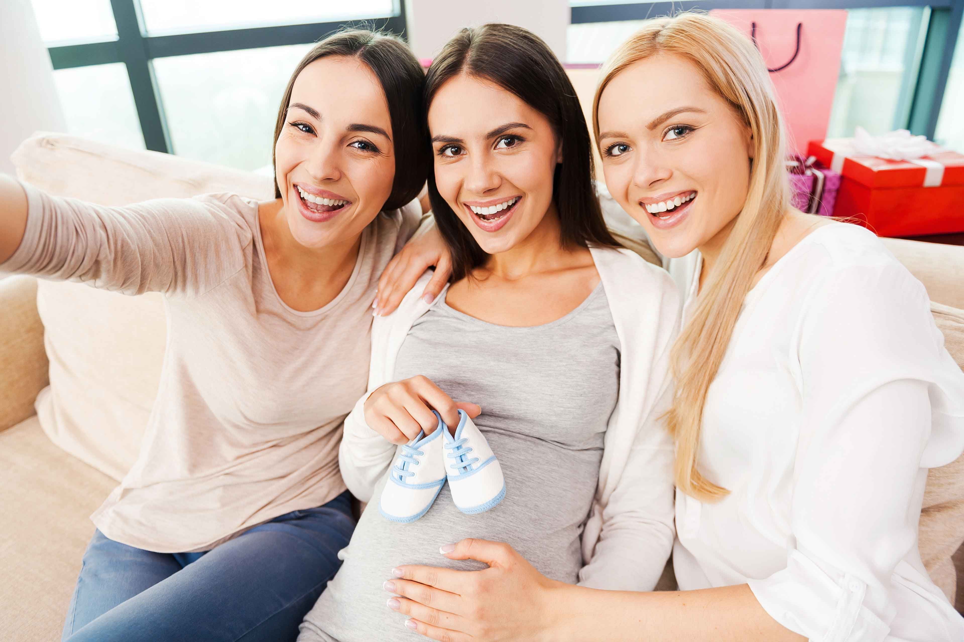Drei Frauen, von denen zwei Babyschuhe in der Hand halten, lächeln und posieren für ein Selfie.