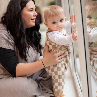 A baby in checkered overalls stands supported at a large glass door, curiously touching their reflection, while an adult kneels nearby, smiling and steadying them. Warm indoor lighting and cozy clothing create a soft, comforting vibe.