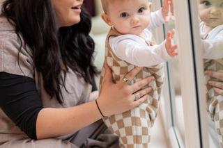 A baby in checkered overalls stands supported at a large glass door, curiously touching their reflection, while an adult kneels nearby, smiling and steadying them. Warm indoor lighting and cozy clothing create a soft, comforting vibe