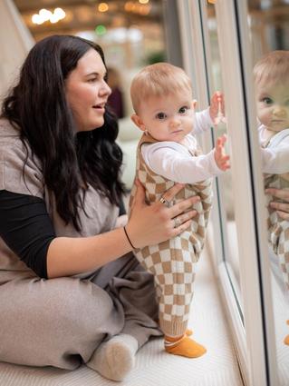 A baby in checkered overalls stands supported at a large glass door, curiously touching their reflection, while an adult kneels nearby, smiling and steadying them. Warm indoor lighting and cozy clothing create a soft, comforting vibe.