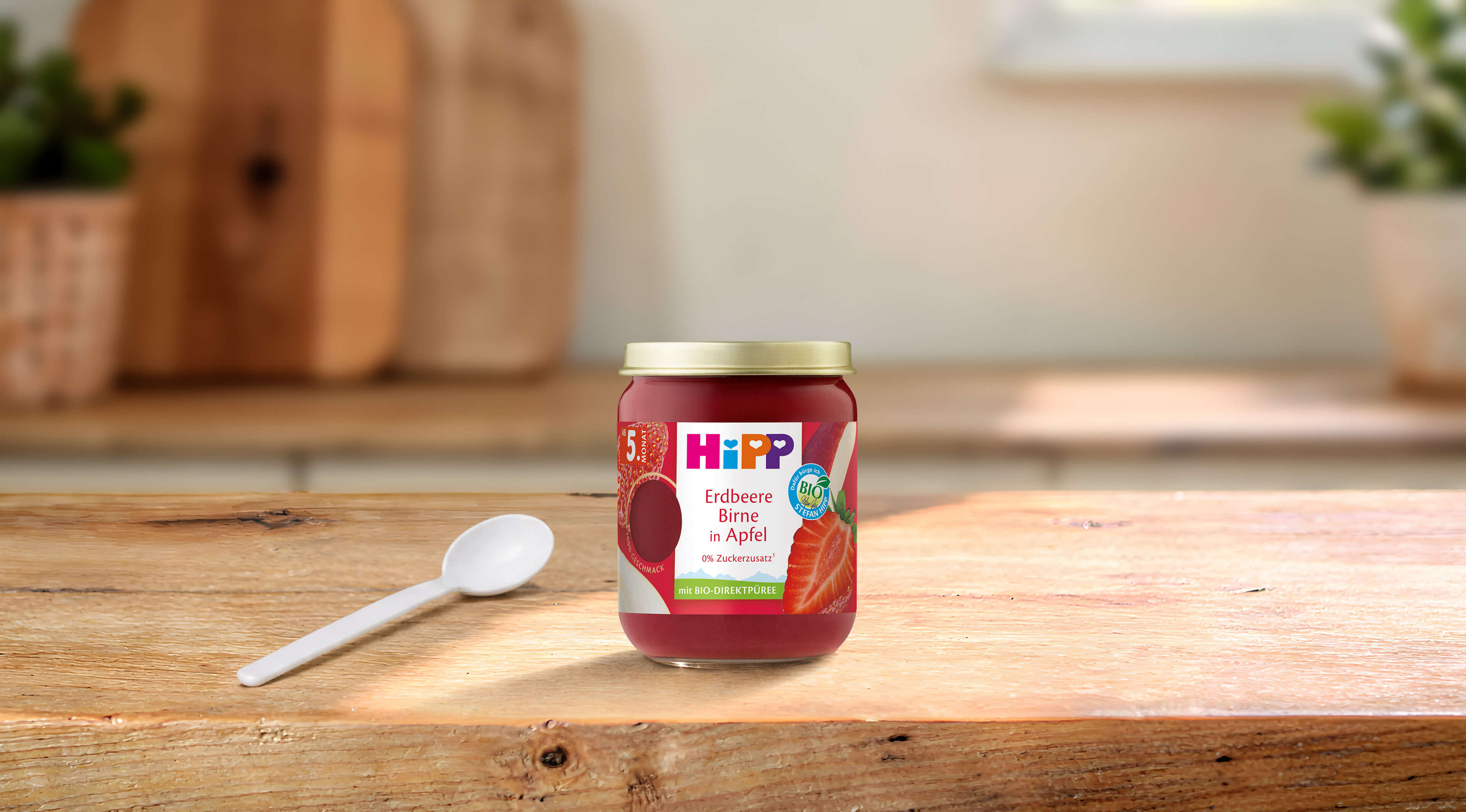 HiPP Bio-Früchteglas Erdbeere Birne in Apfel auf Holztisch, daneben ein weißer Babylöffel in heller Küchenumgebung