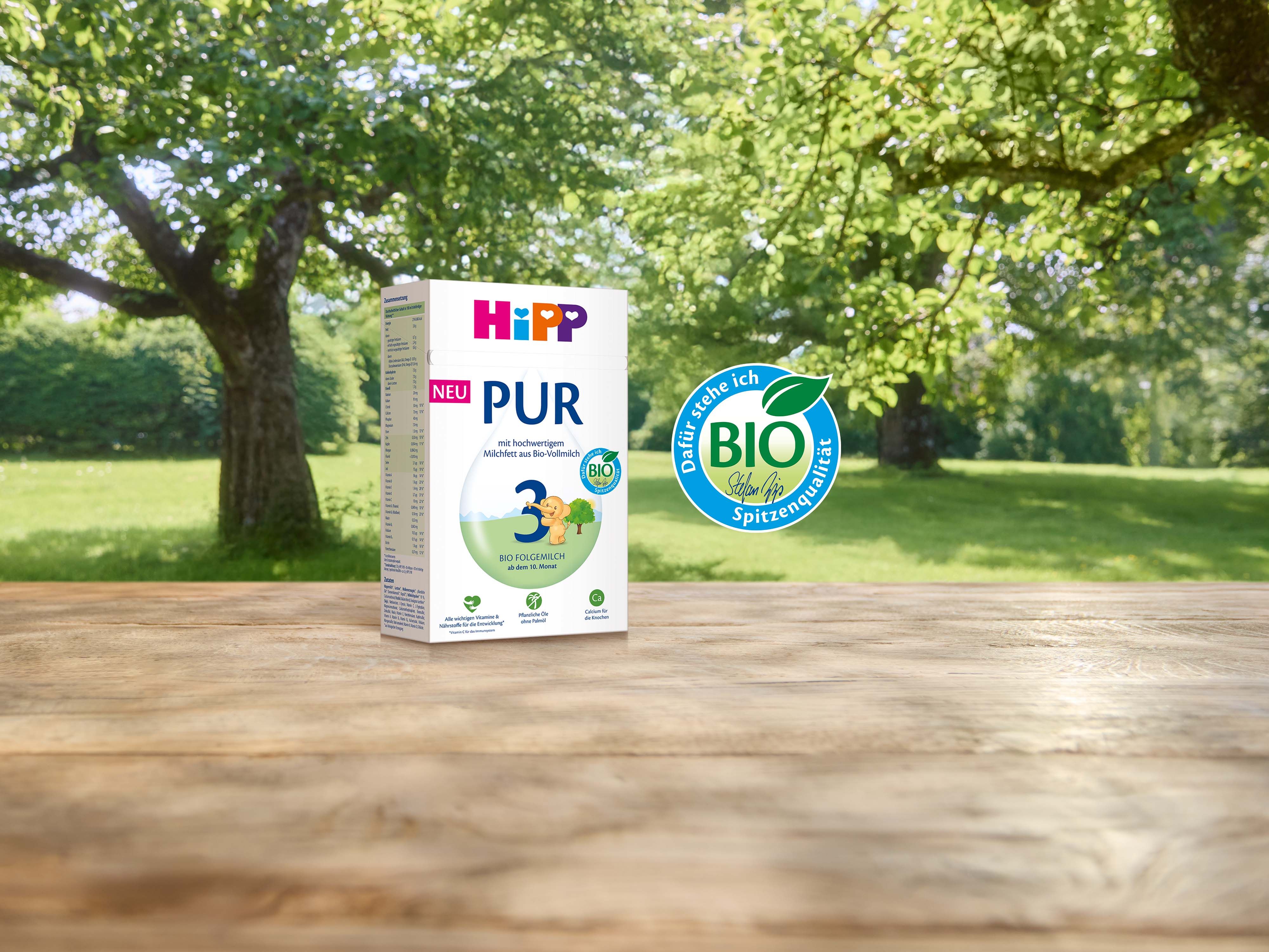 Packung HiPP PUR Bio-Babynahrung auf Holztisch vor grüner Baumkulisse