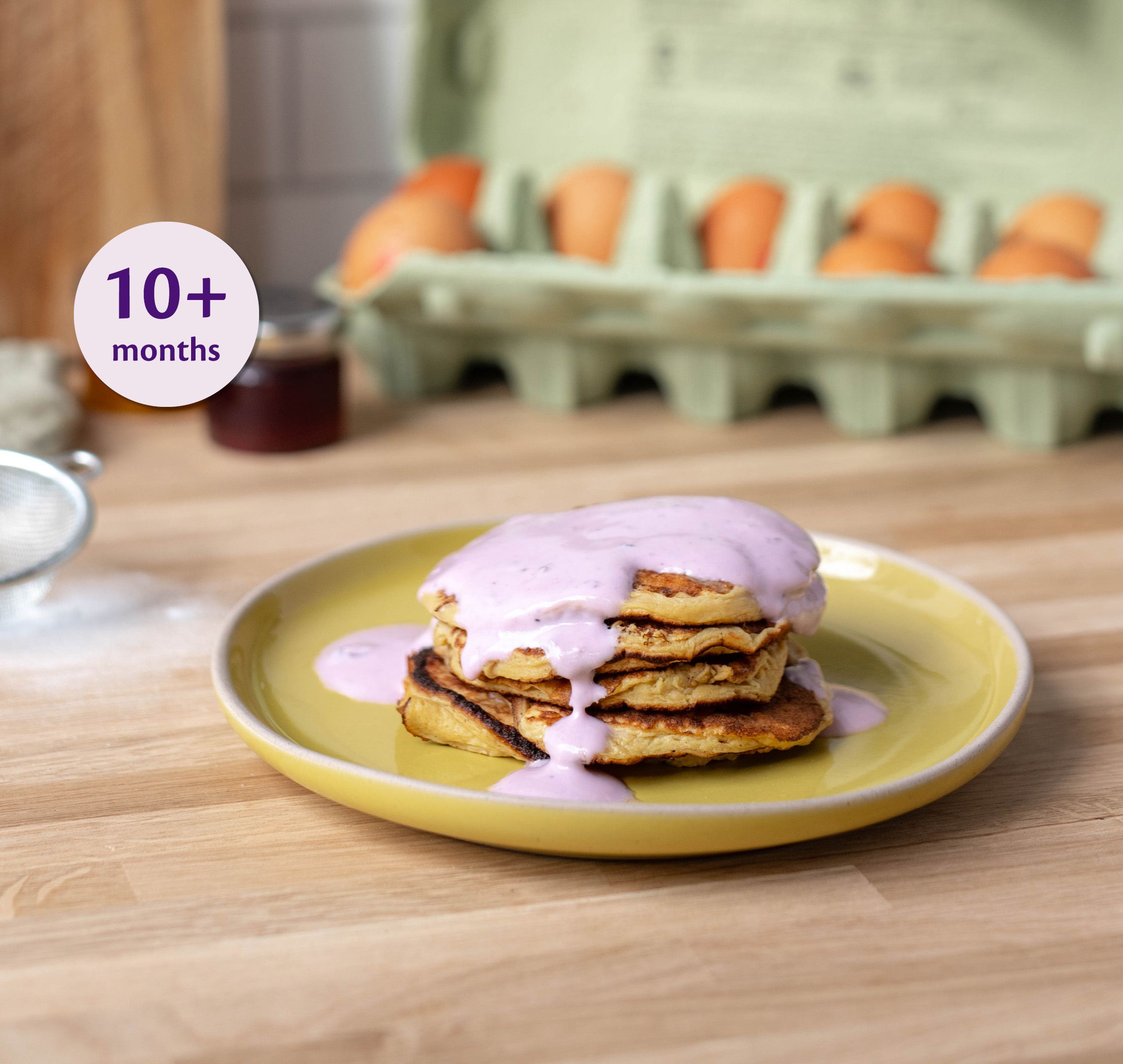 Stack of mini apple pancakes on a yellow plate topped with pink yogurt, labelled suitable for babies 10+ months.