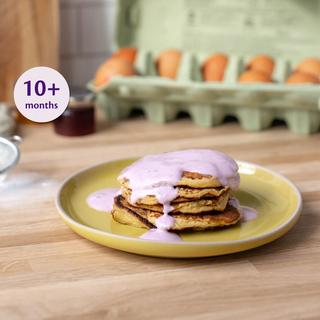 Stack of mini apple pancakes on a yellow plate topped with pink yogurt, labelled suitable for babies 10+ months.