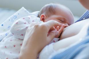 Ein Baby, das von seiner Mutter gestillt wird, wobei das Gesicht des Babys gut sichtbar ist und die Hand der Mutter den Kopf des Babys sanft berührt.