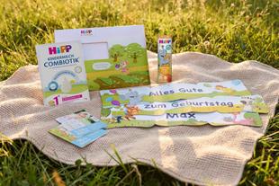 Kinderpaket mit HiPP Kindermilch Combiotik, Geburtstagsbanner mit Aufschrift 'Alles Gute zum Geburtstag Max', Geschenkbox mit Tierillustrationen und Rabatt-Coupons auf Decke in einer Wiese