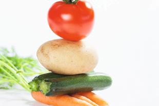 Ein Stapel frisches Gemüse, darunter eine Tomate, eine Kartoffel, eine Zucchini und eine Karotte, mit einem Hauch von grünen Kräutern im Hintergrund.