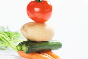 Ein Stapel frisches Gemüse, darunter eine Tomate, eine Kartoffel, eine Zucchini und eine Karotte, mit einem Hauch von grünen Kräutern im Hintergrund.