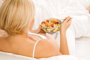 Eine Frau mit blondem Haar, die einen weißen Schlafanzug trägt und eine Schüssel Müsli mit Erdbeeren und Blaubeeren isst.