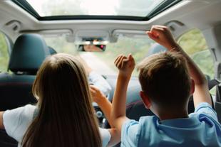 Zwei Kinder, ein Mädchen und ein Junge, sitzen mit erhobenen Händen in einem Auto und scheinen eine Fahrt zu genießen.