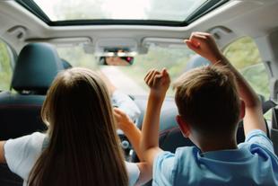 Zwei Kinder, ein Mädchen und ein Junge, sitzen mit erhobenen Händen in einem Auto und scheinen eine Fahrt zu genießen.