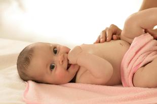 Ein auf einem rosa Handtuch liegendes Baby, das mit einer Hand sanft seinen Bauch berührt und dabei einen freudigen Ausdruck zeigt.