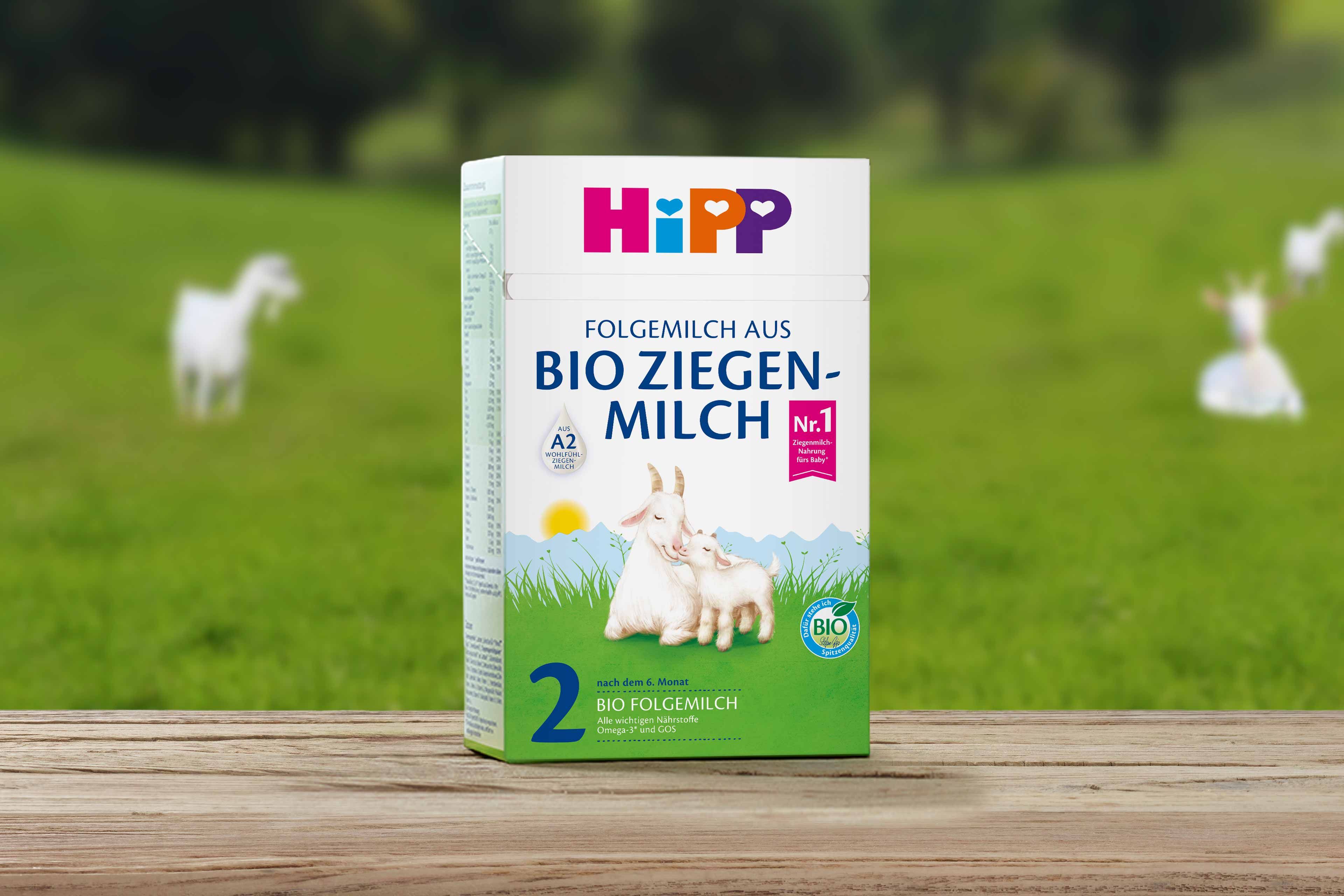 Packung HiPP Folgemilch aus Bio-Ziegenmilch 2 auf Holztisch vor grüner Wiese mit Ziegen im Hintergrund.