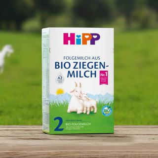 Packung HiPP Folgemilch aus Bio-Ziegenmilch 2 auf Holztisch vor grüner Wiese mit Ziegen im Hintergrund.