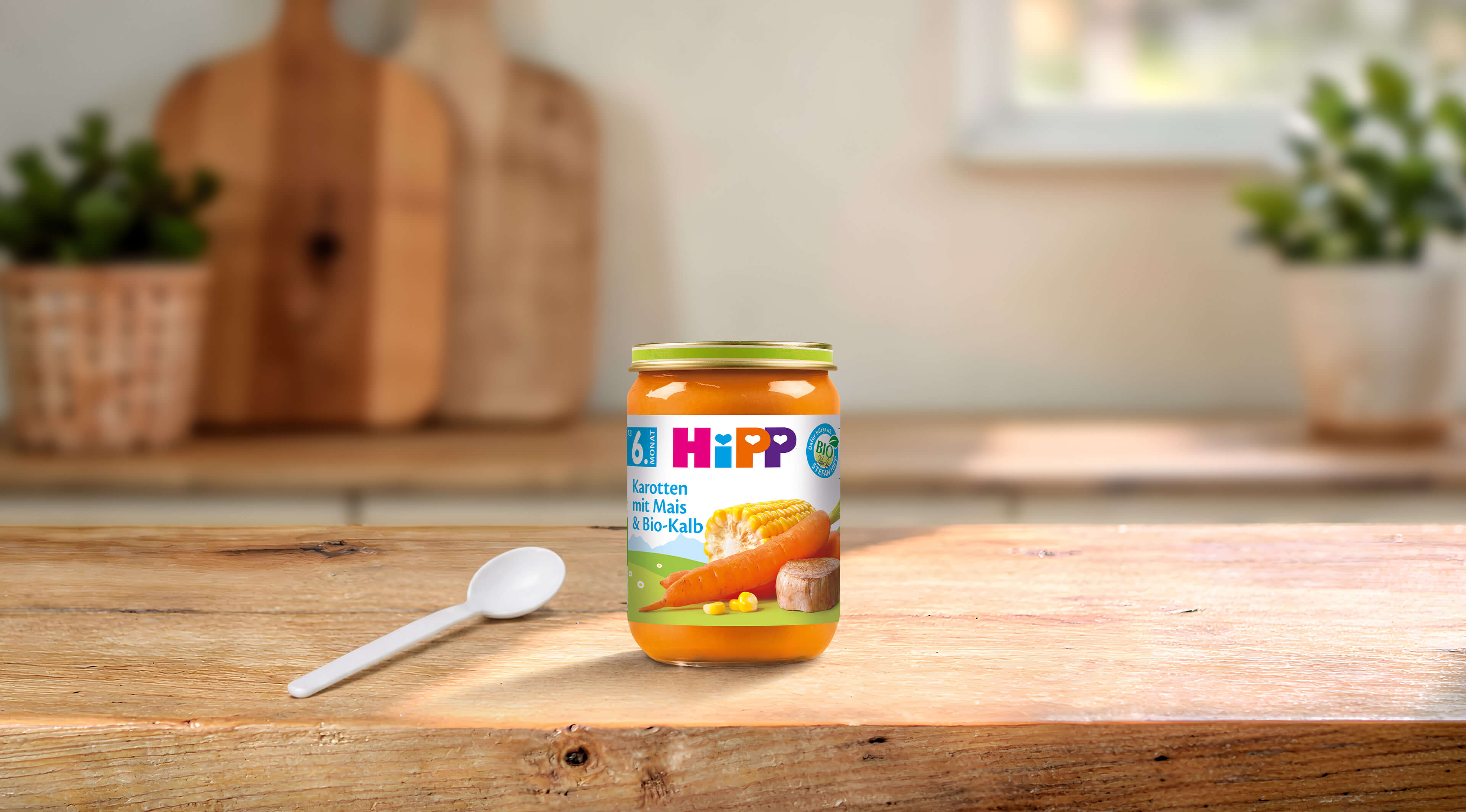 HiPP Bio-Gläschen Karotten mit Mais & Bio-Kalb ab 6. Monat auf Holztisch mit Babylöffel in heller Küche
