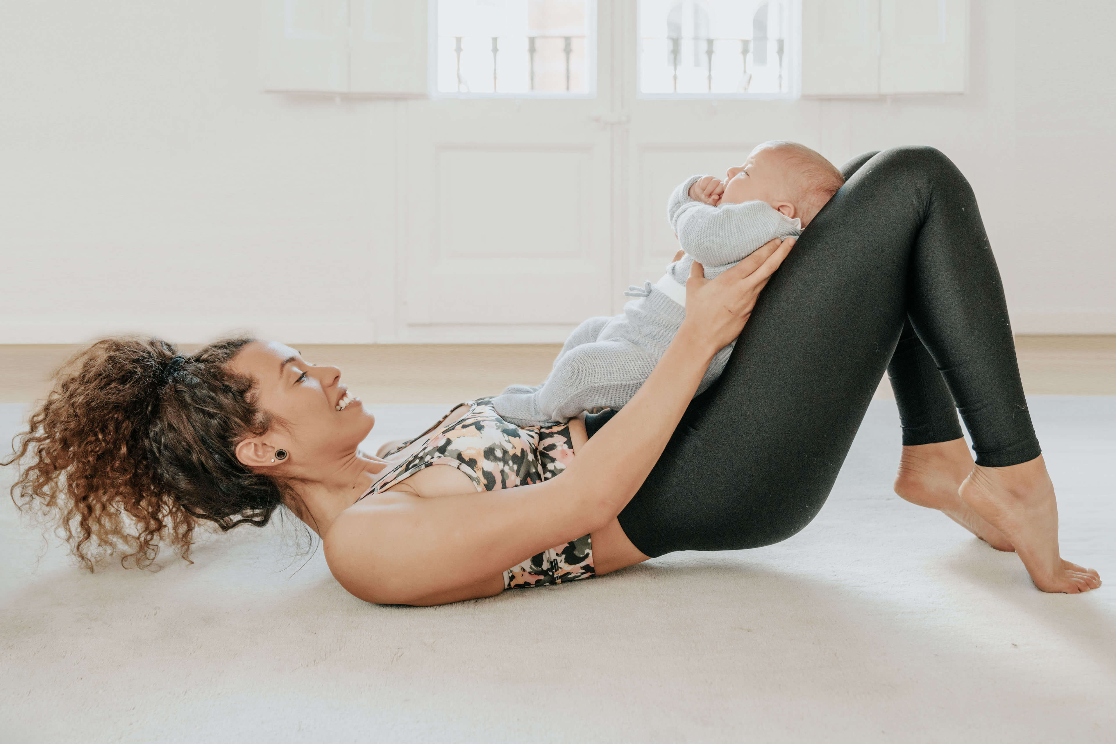 Eine auf dem Boden liegende Frau mit einem Baby im Arm, in entspannter Pose und mit einem freudigen Gesichtsausdruck.