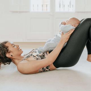 Eine auf dem Boden liegende Frau mit einem Baby im Arm, in entspannter Pose und mit einem freudigen Gesichtsausdruck.