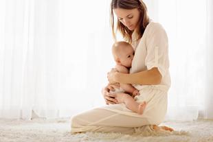Eine Frau in einem weißen Kleid, die ein Baby an sich drückt, beide auf einer weichen Unterlage sitzend, in natürliches Licht getaucht.