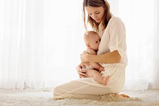Eine Frau in einem weißen Kleid, die ein Baby an sich drückt, beide auf einer weichen Unterlage sitzend, in natürliches Licht getaucht.