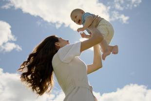 Frau mit langen braunen Haaren hebt Baby in die Luft vor blauem Himmel mit Wolken