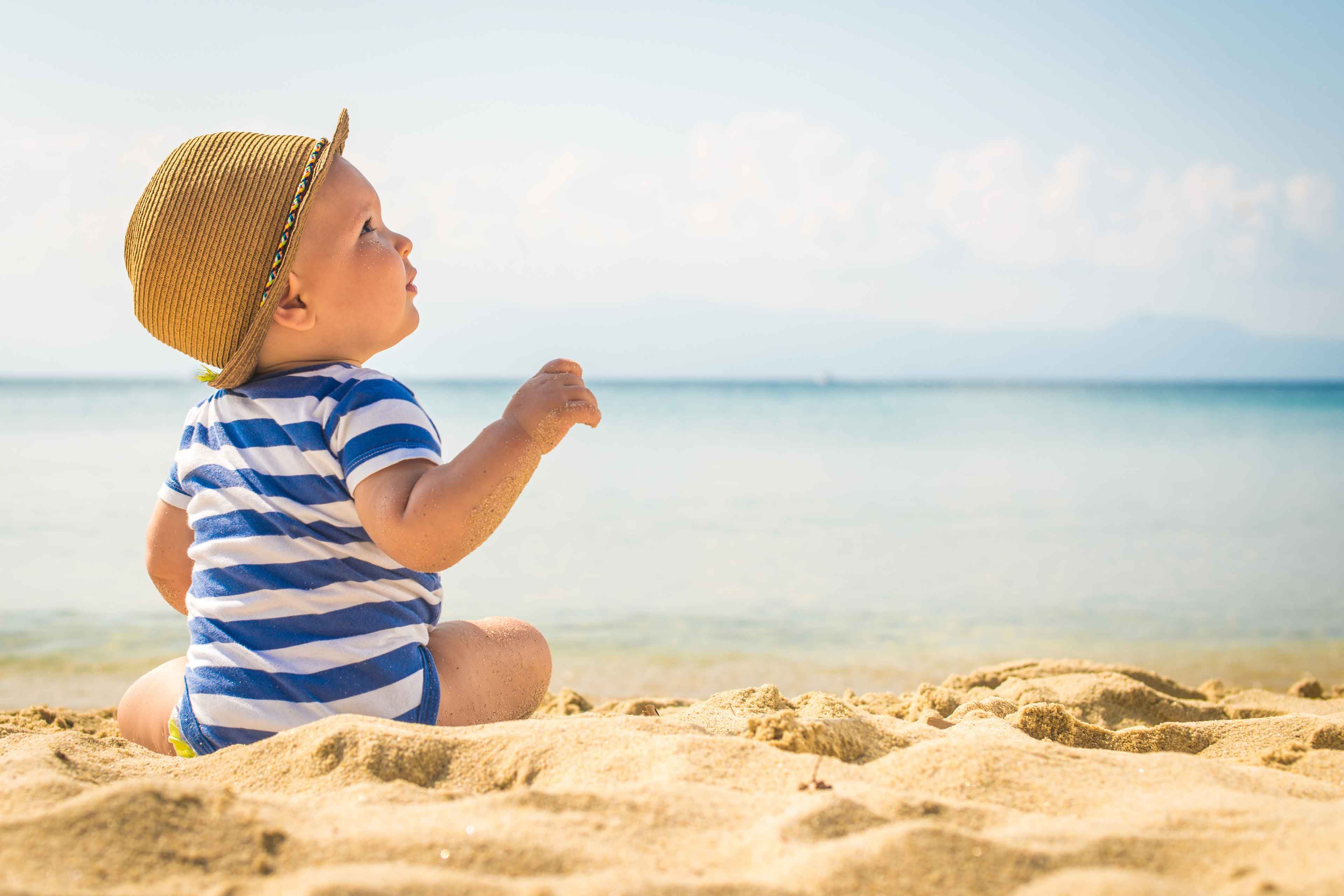 Ein kleines Kind in einem gestreiften Outfit und mit einem Hut sitzt an einem Sandstrand und blickt in die Ferne mit einer ruhigen Kulisse des Meeres und der fernen Berge.
