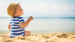 Ein kleines Kind in einem gestreiften Outfit und mit einem Hut sitzt an einem Sandstrand und blickt in die Ferne mit einer ruhigen Kulisse des Meeres und der fernen Berge.