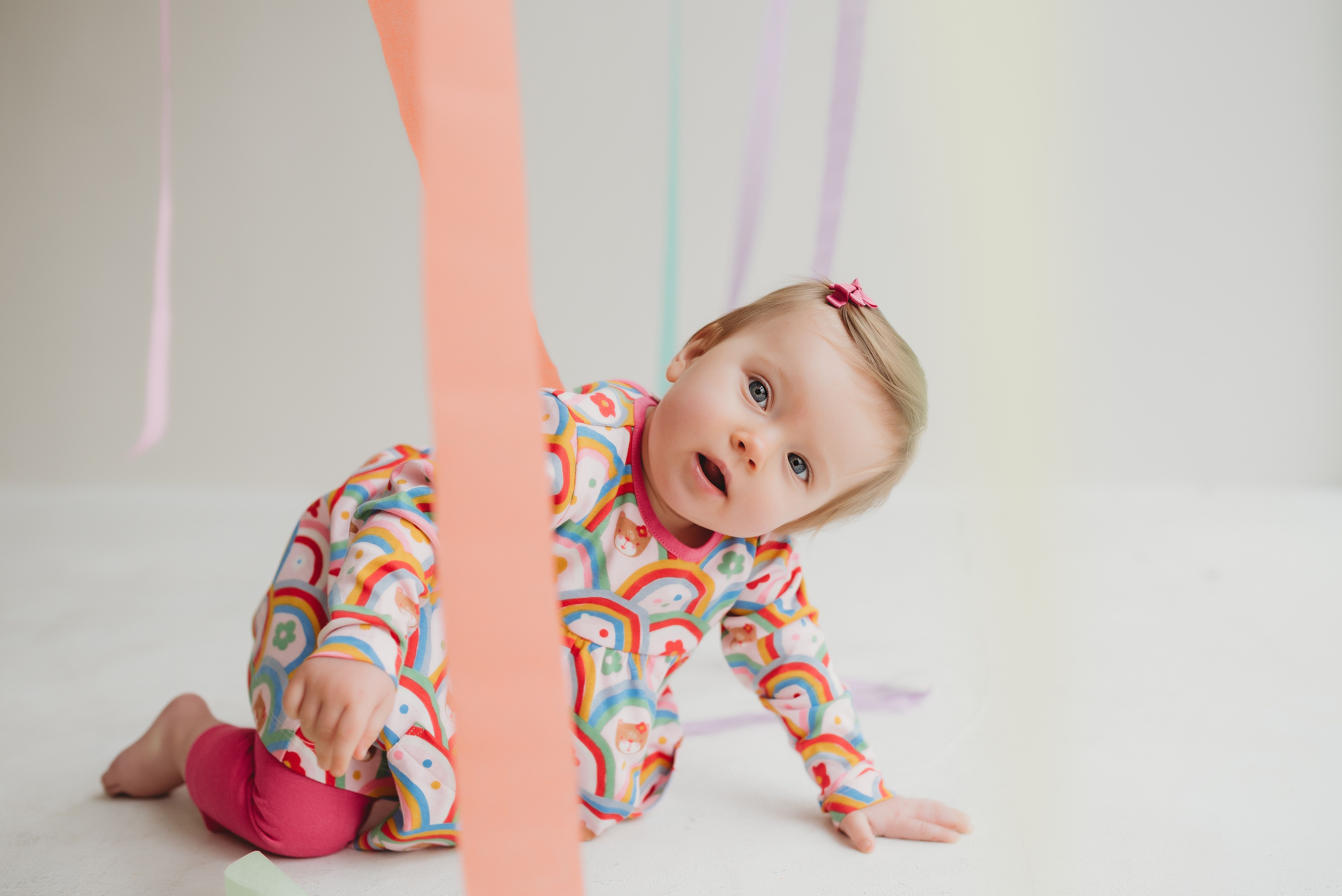 Smiling baby in a colourful rainbow-print outfit playing on the floor, promoting up to 40% off Lilly + Sid baby clothes.