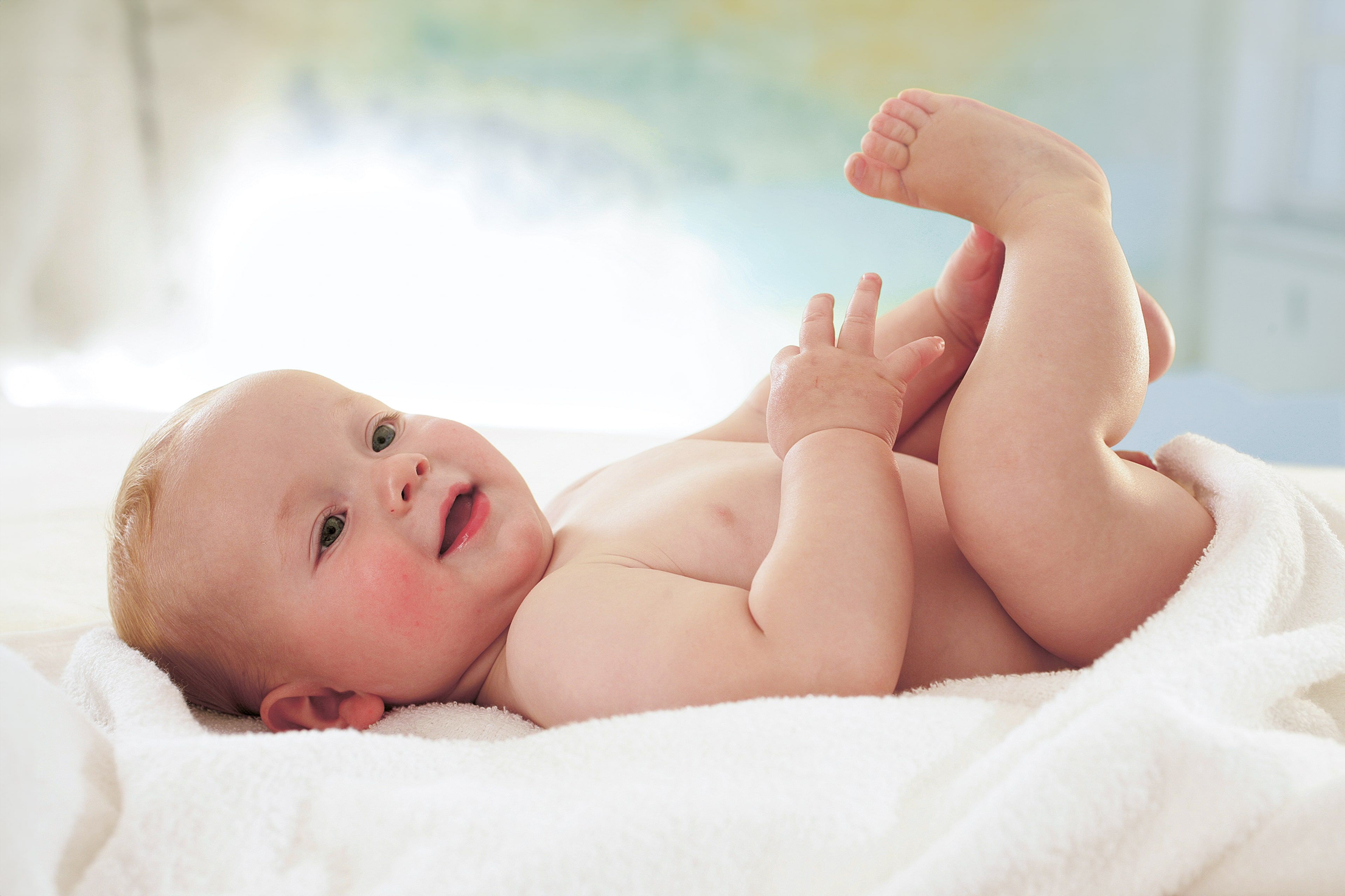 Ein fröhliches Baby, das auf einer weißen Fläche liegt, mit seinen Füßen spielt und mit einem strahlenden Gesichtsausdruck nach oben schaut.