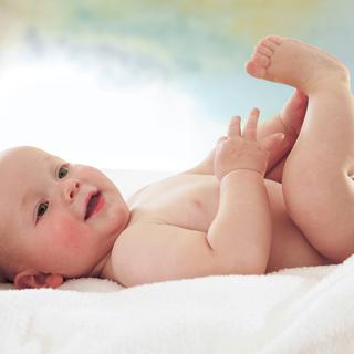 Ein fröhliches Baby, das auf einer weißen Fläche liegt, mit seinen Füßen spielt und mit einem strahlenden Gesichtsausdruck nach oben schaut.