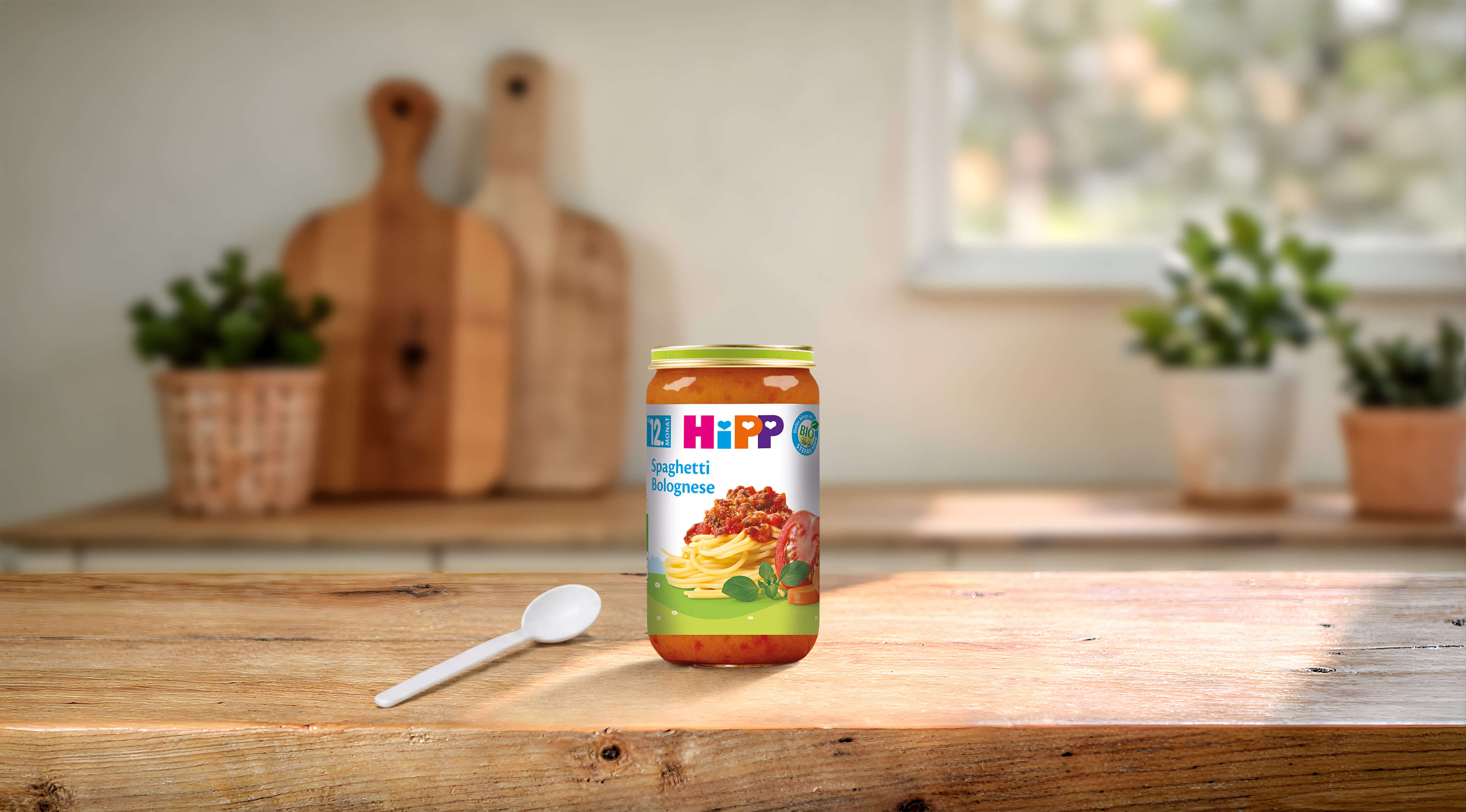 HiPP Bio-Glas Spaghetti Bolognese 250g ab 12 Monaten auf Holztisch mit Löffel in heller Küchenumgebung