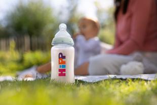Eine HiPP Milchflasche steht im Gras, im Hintergrund sieht man verschwommen ein Baby und eine Frau