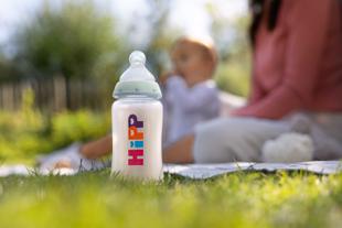 Eine HiPP Milchflasche steht im Gras, im Hintergrund sieht man verschwommen ein Baby und eine Frau