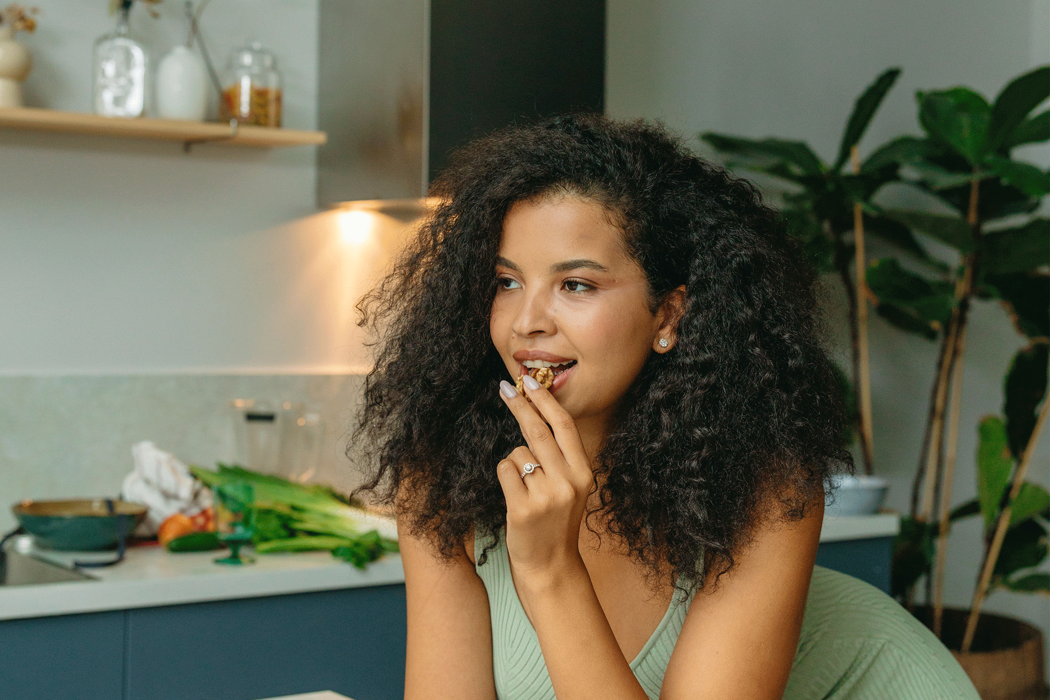 A woman with long curly dark hair and a light green tank top standing in a kitchen and eating a cluster of nuts or granola while looking off to the side.
