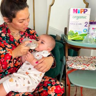 A mother in colourful pyjamas bottle-feeding her baby in a living room, with HiPP Organic Stage 2 formula visible on a nearby stand.