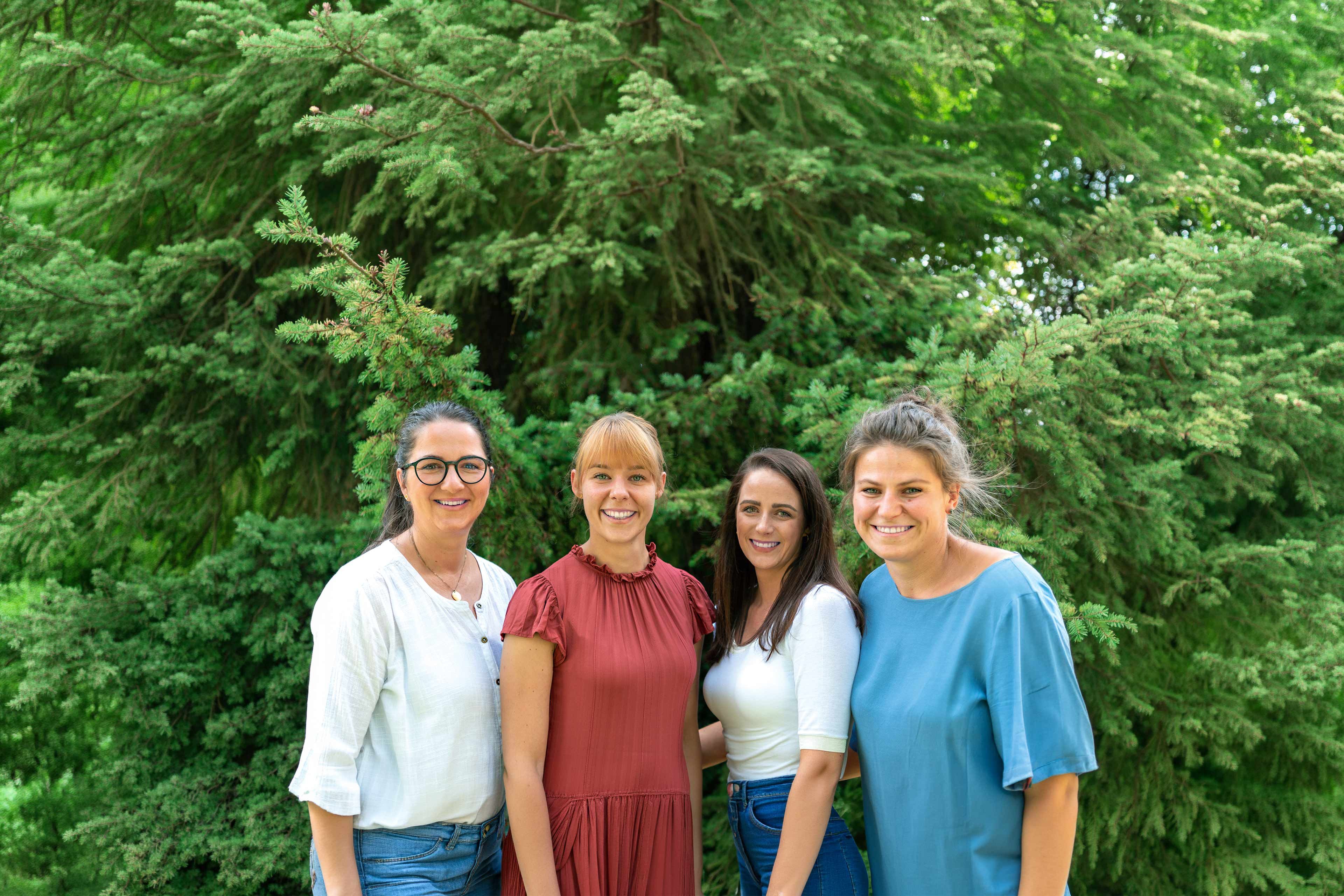 Vier Frauen stehen nebeneinander vor einem großen Nadelbaum und lächeln in die Kamera.