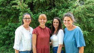 Vier Frauen stehen nebeneinander vor grünen Bäumen und lächeln in die Kamera