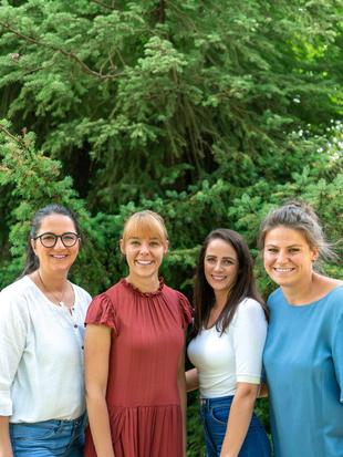 Vier Frauen stehen nebeneinander vor einem großen Nadelbaum und lächeln in die Kamera.