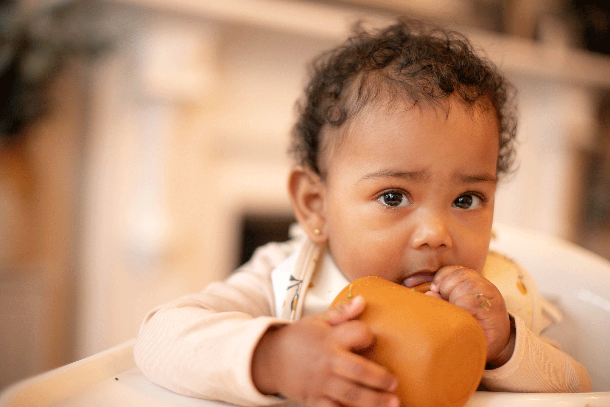 Close-up of a young toddler with curly dark hair sitting in a white high chair wearing a patterned bib, holding a small mustard-orange silicone beaker with both hands while looking toward the camera.
