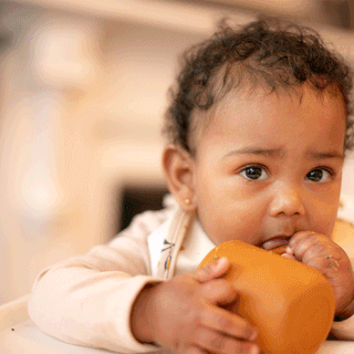 Close-up of a young toddler with curly dark hair sitting in a white high chair wearing a patterned bib, holding a small mustard-orange silicone beaker with both hands while looking toward the camera.