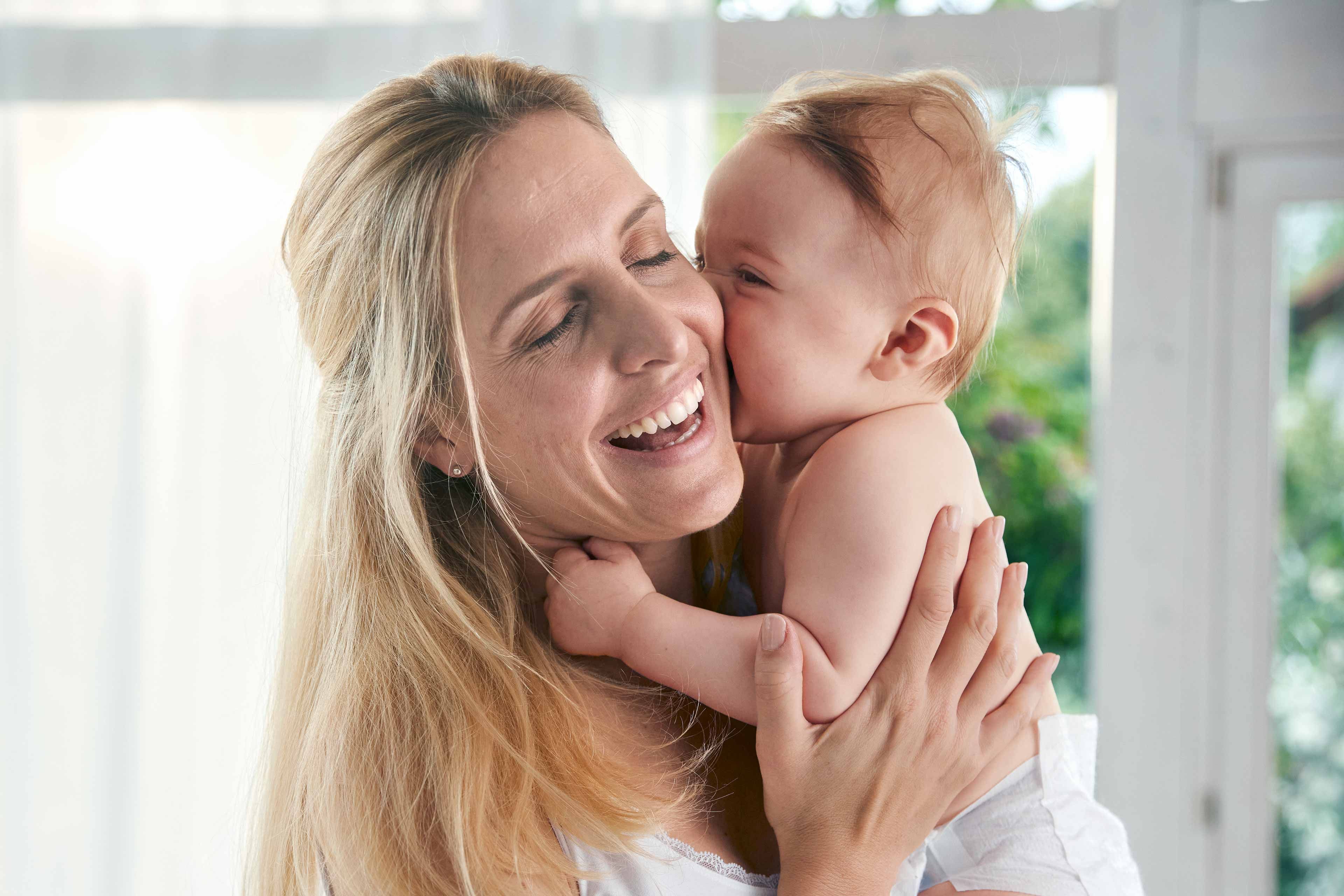 Ein zärtlicher Moment zwischen einer Mutter und ihrem Baby, in dem die Mutter ihr Baby freudig an ihr Gesicht drückt.