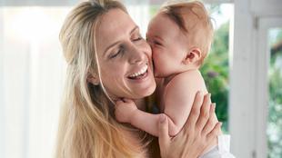 Ein zärtlicher Moment zwischen einer Mutter und ihrem Baby, in dem die Mutter ihr Baby freudig an ihr Gesicht drückt.