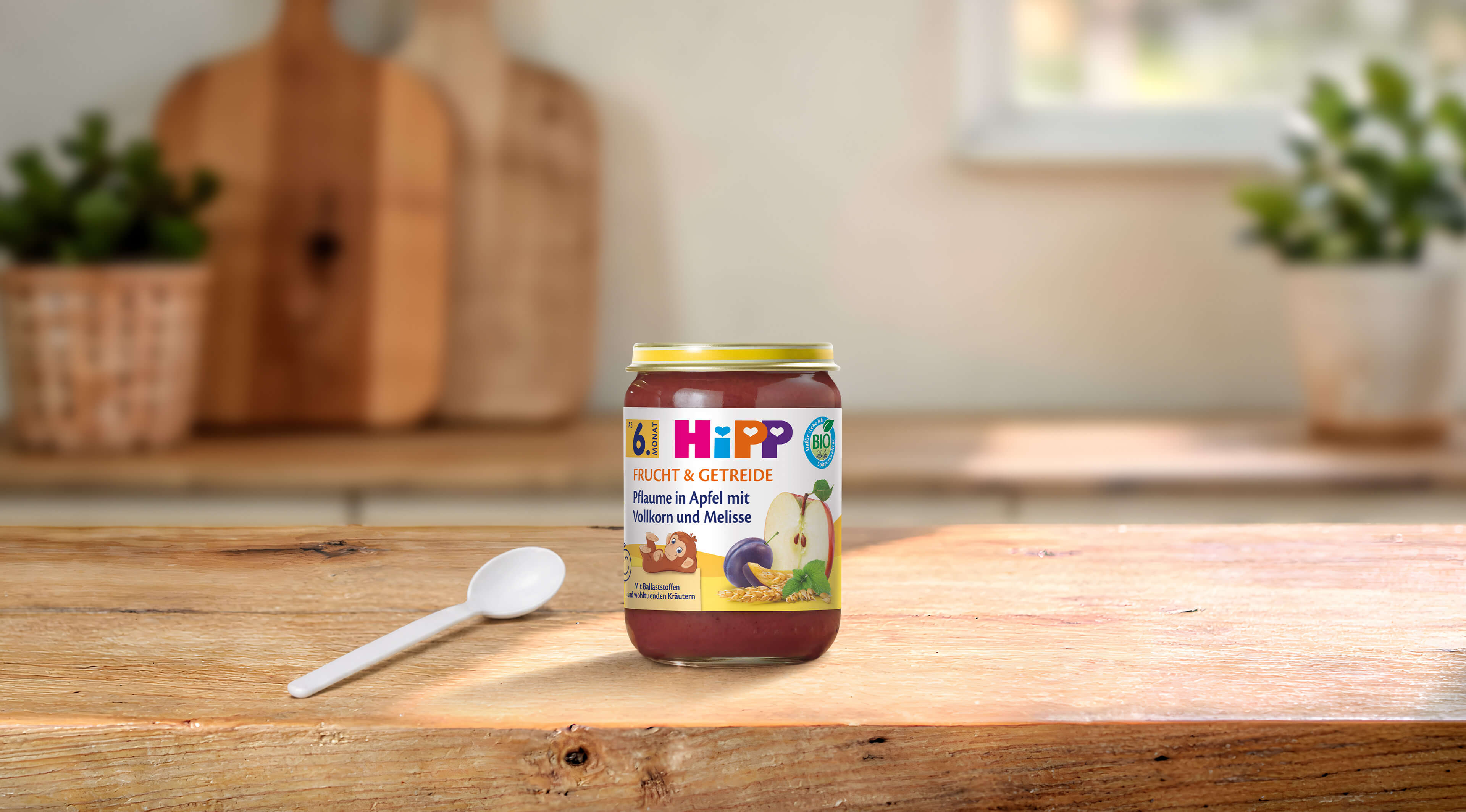 HiPP Bio-Gläschen Pflaume in Apfel mit Vollkorn und Melisse auf Holztisch mit Löffel in heller Küchenumgebung