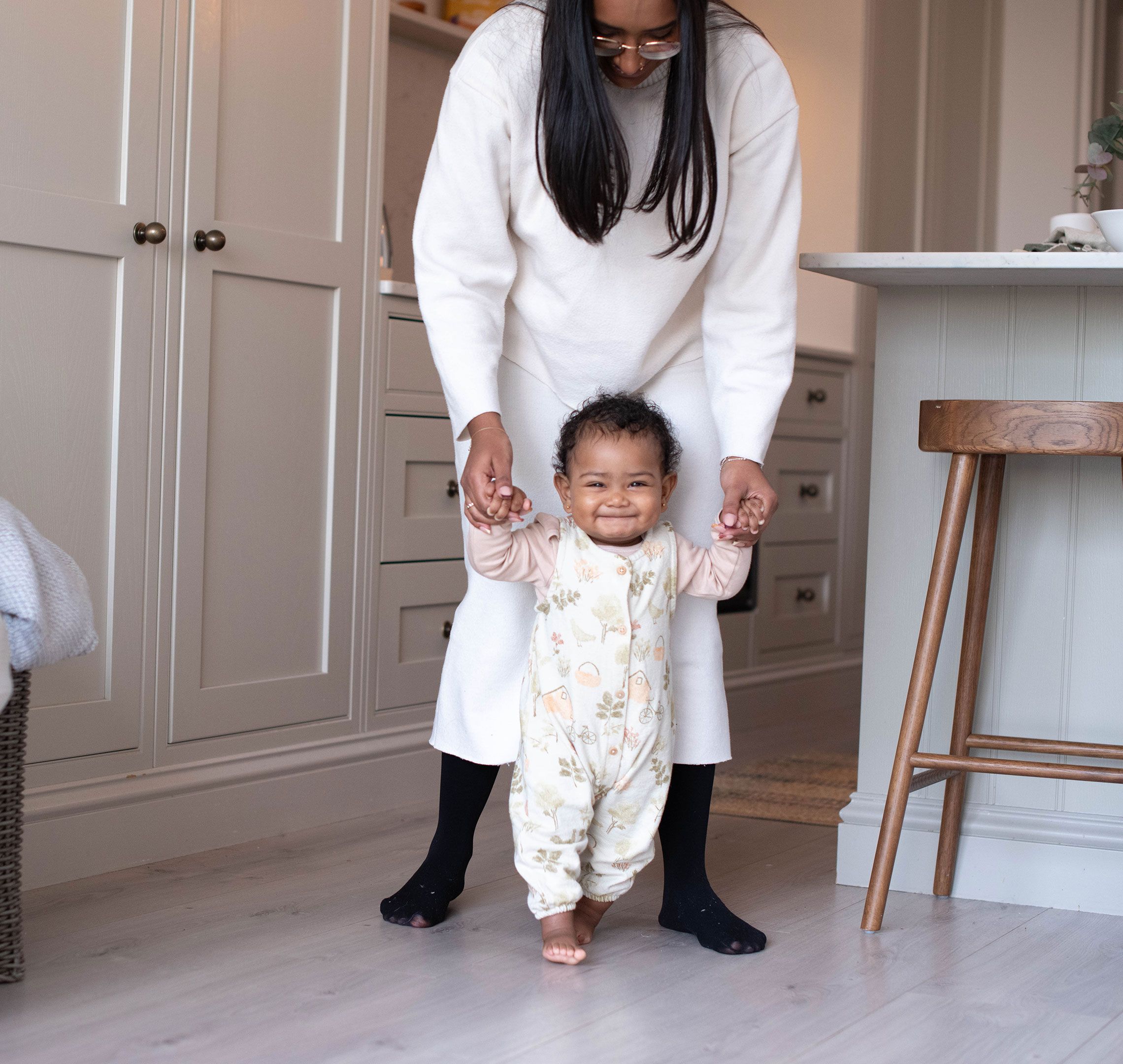Smiling baby taking early steps indoors while a parent gently supports their hands from behind