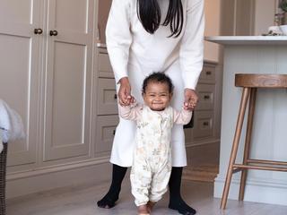 Smiling baby taking early steps indoors while a parent gently supports their hands from behind