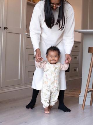 Smiling baby taking early steps indoors while a parent gently supports their hands from behind