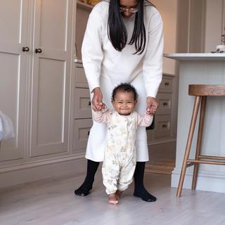Smiling baby taking early steps indoors while a parent gently supports their hands from behind