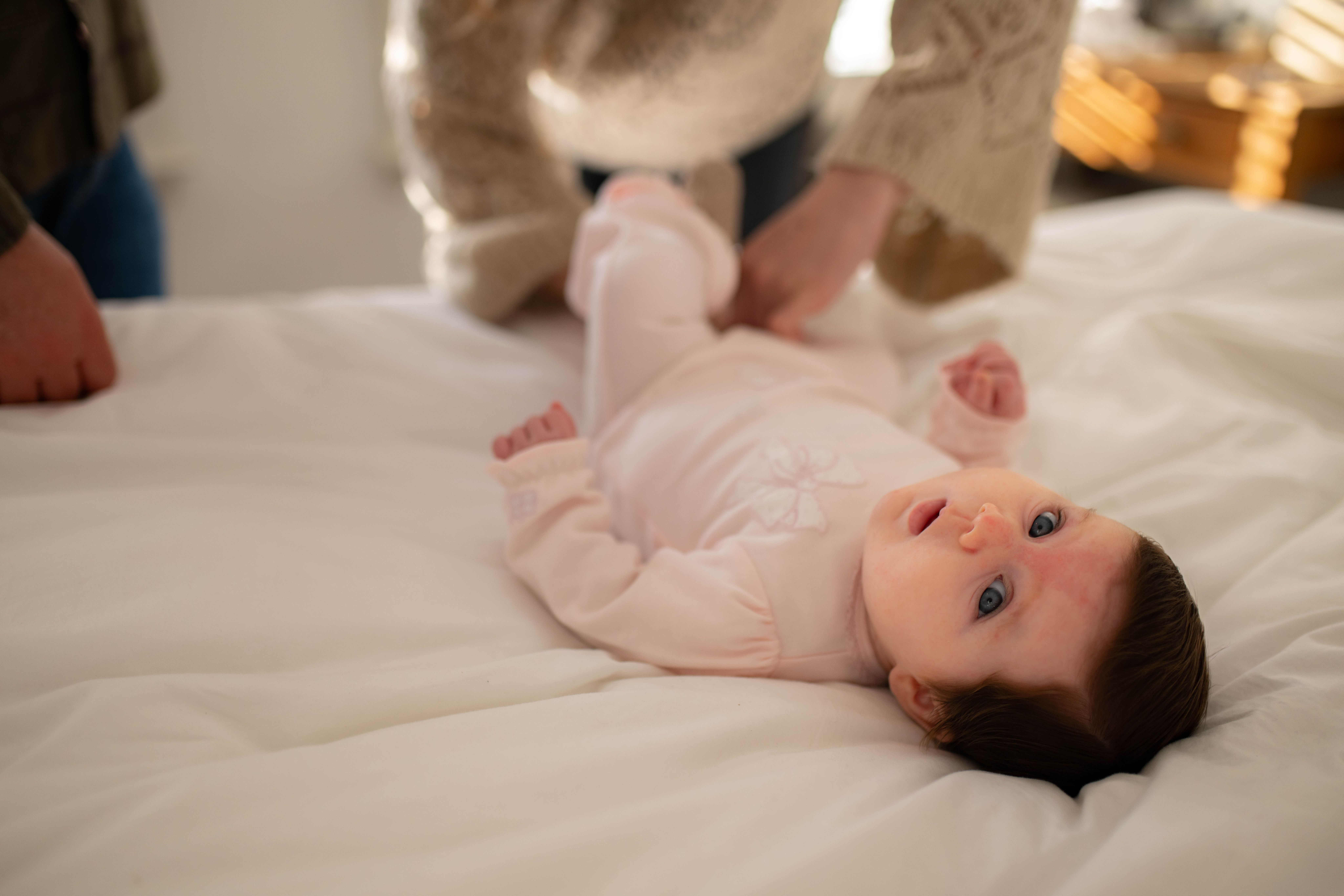 Young baby lying on a bed during a nappy change, looking up while a parent gently tends to them.