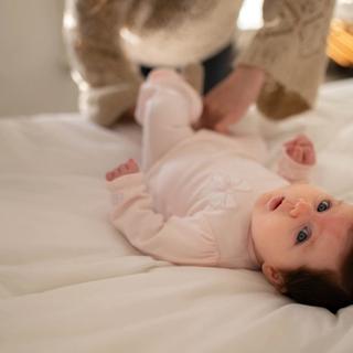 Young baby lying on a bed during a nappy change, looking up while a parent gently tends to them.