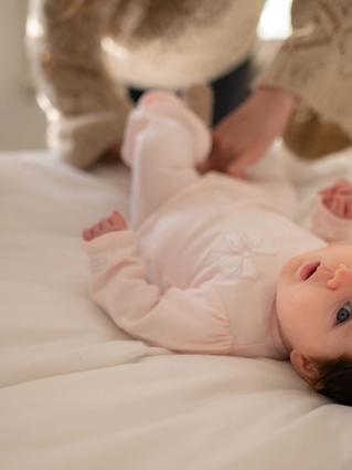 Young baby lying on a bed during a nappy change, looking up while a parent gently tends to them.